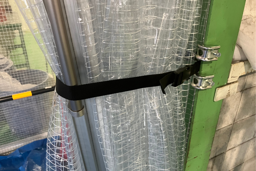 糸入り透明ビニールカーテンをバックル付きのベルトで柱に束ねて固定した状態。作業時の動線を確保するための、実用的で機能的なカーテン収納の施工事例です。