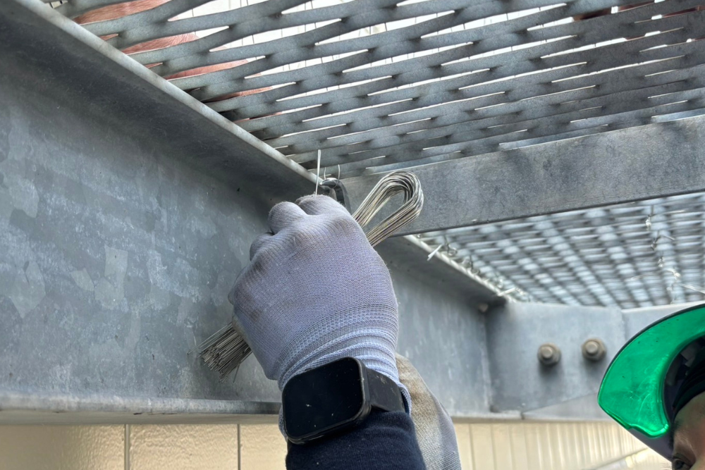 鳥害対策の施工風景。作業員が手作業で防鳥ピンを足場のメッシュ部分に固定している様子。細部まで丁寧な施工を行うことで、鳥の飛来を長期的に防止します。