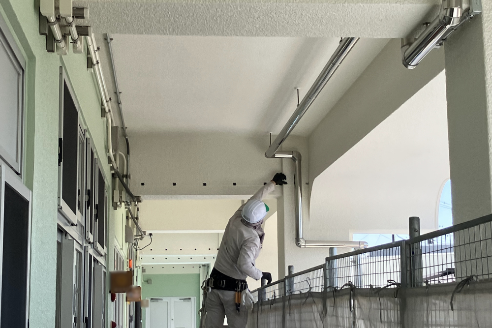 屋外通路の梁の部分に、防鳥ネット固定用具を等間隔で取り付けている施工中の様子。