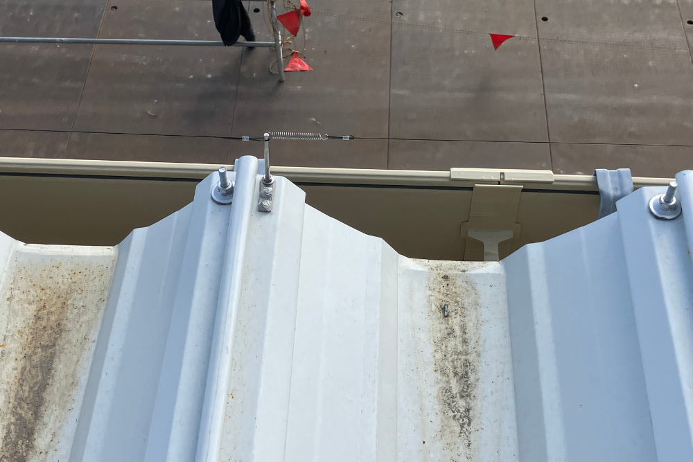 工場の折板屋根の軒先に設置された防鳥ワイヤーを真上から撮影。雨樋付近の鳥の飛来を防ぐため、専用の支柱とスプリングでワイヤーが固定されています。