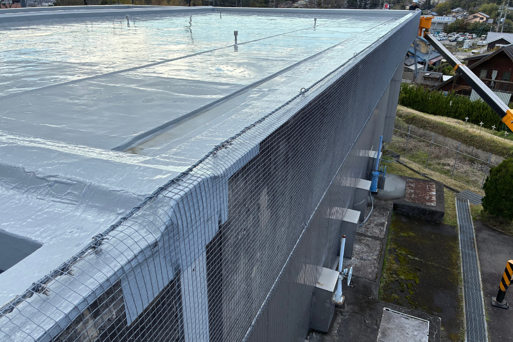 防水処理が施された建物の平らな屋上と、その縁から外壁へと続くグレーの防護ネットの設置状況