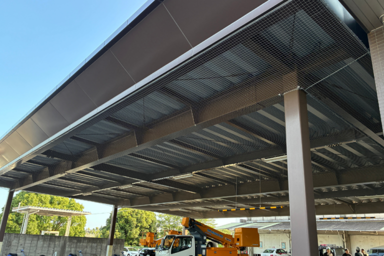 高所作業車を用いた大型施設の庇下への施工風景。広範囲の天井部分全体に鳥よけネットを綺麗に展張している様子