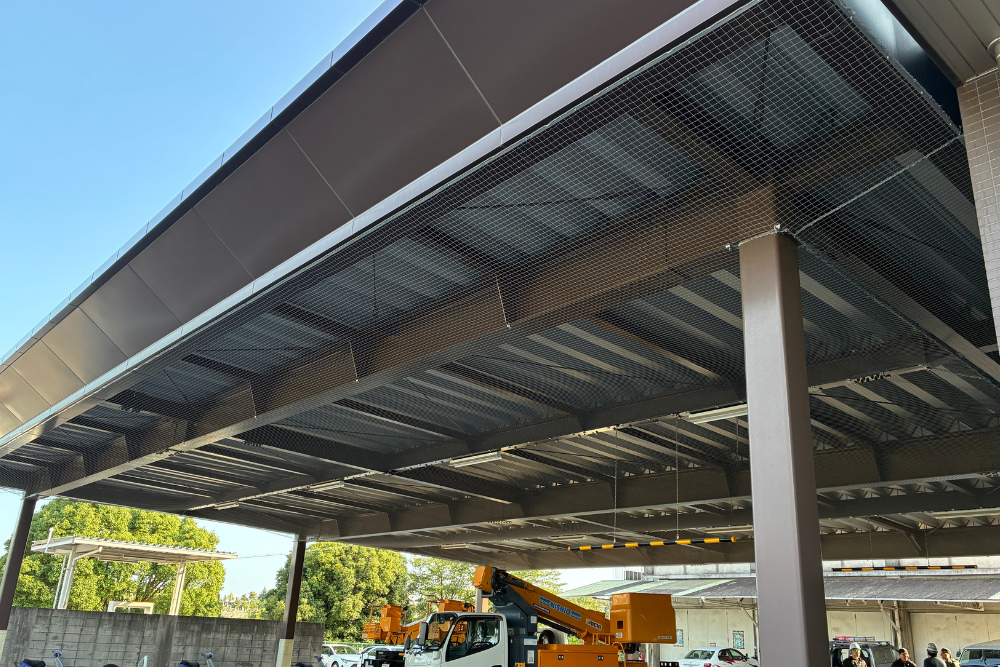 高所作業車を用いた大型施設の庇下への施工風景。広範囲の天井部分全体に鳥よけネットを綺麗に展張している様子