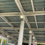 高速道路料金所の屋根裏構造を見上げた様子。白い鉄骨が組まれた天井には鳥よけネットが張られ、柱の上部に設置された照明が点灯して周囲を照らしています。
