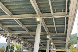 高速道路料金所の屋根裏構造を見上げた様子。白い鉄骨が組まれた天井には鳥よけネットが張られ、柱の上部に設置された照明が点灯して周囲を照らしています。
