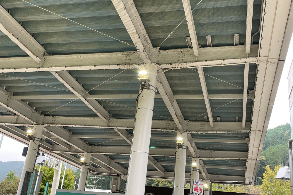 高速道路料金所の屋根裏構造を見上げた様子。白い鉄骨が組まれた天井には鳥よけネットが張られ、柱の上部に設置された照明が点灯して周囲を照らしています。