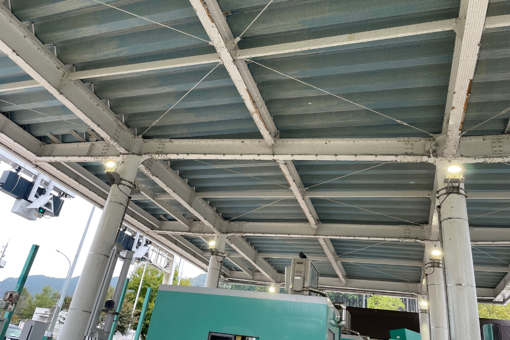 白い鉄骨で組まれた高速道路料金所の天井部分。鉄骨全体を覆うように鳥よけネットが張られ、下には緑色の料金所ブースやETC設備が見えます。