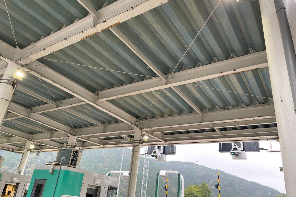 高速道路料金所の屋根を下から見上げた様子。白い鉄骨の天井全面に鳥よけネットが張られ、下部には緑色の料金所ブースやETC設備が設置されています。
