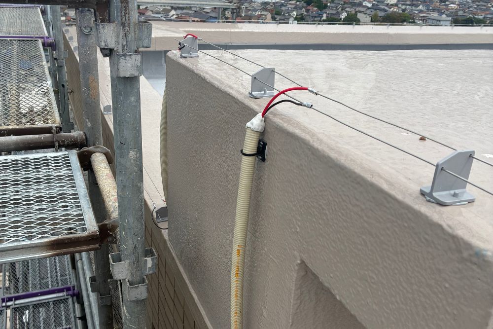 屋上の縁に設置された鳥よけ電気ショックワイヤーの近影。足場が組まれた施工現場で、碍子と赤黒の配線が接続されている様子です。