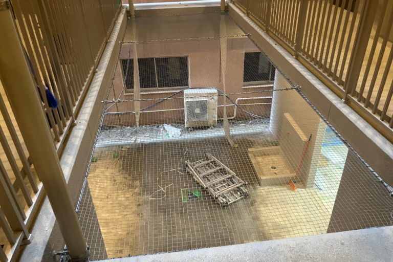 上部から見下ろしたアングルで撮影された、マンションの吹き抜け空間に設置された落下防止対策用ネットの施工風景。